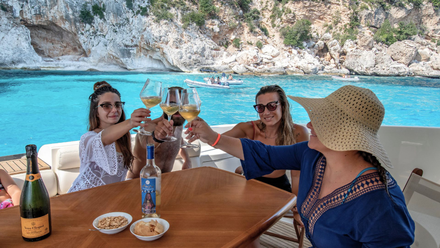 Gruppo di persone sedute a un tavolo su uno yacht di lusso in mare aperto, con vista su una baia nascosta e scogliere rocciose. Una bottiglia di champagne e bicchieri sono sul tavolo.