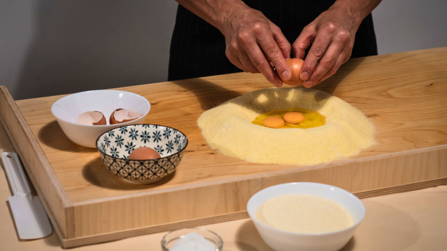 Persona che prepara ravioli tradizionali su un tagliere di legno con ingredienti freschi. Due ciotole con uova e un piatto di farina