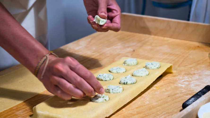 Una persona prepara ravioli con ripieno di formaggio su un tavolo di legno, circondata da altri ravioli già pronti. L'immagine raffigura un'attività di laboratorio di cucina.