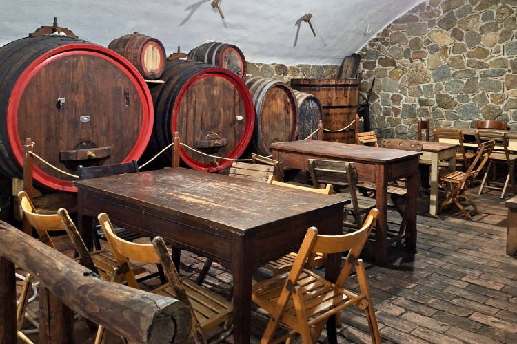 Interno di una cantina con tavoli e sedie, e grandi botti di legno per la produzione del vino.