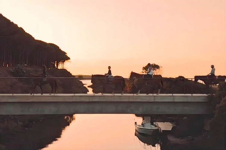 Tre persone a cavallo attraversano un ponte su un fiume al tramonto
