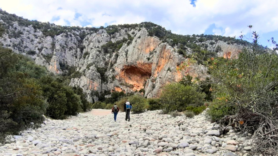 Due escursionisti camminano su un sentiero sassoso tra rocce calcaree e macchia mediterranea, diretti verso Cala Luna, con vista sul mare turchese