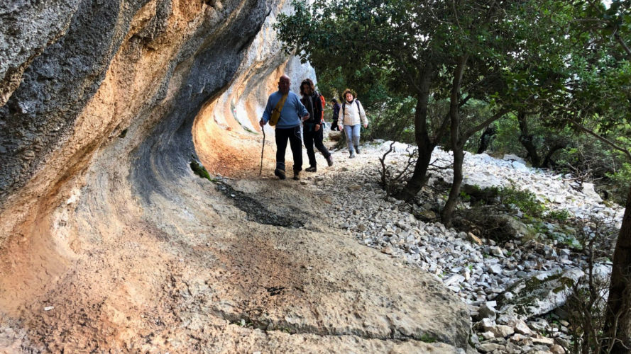Gente caminando por un camino de tierra entre rocas y vegetación mediterránea.