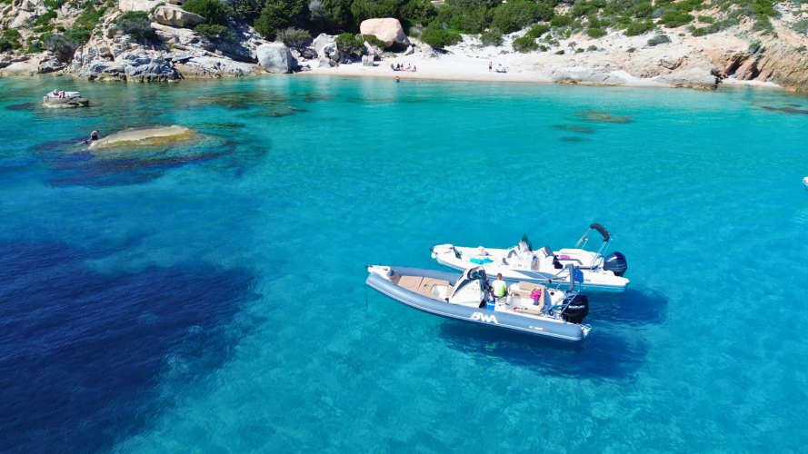 Immagine di un'area marina con barche ormeggiate e persone sulla spiaggia, acque turchesi e paesaggi incontaminati