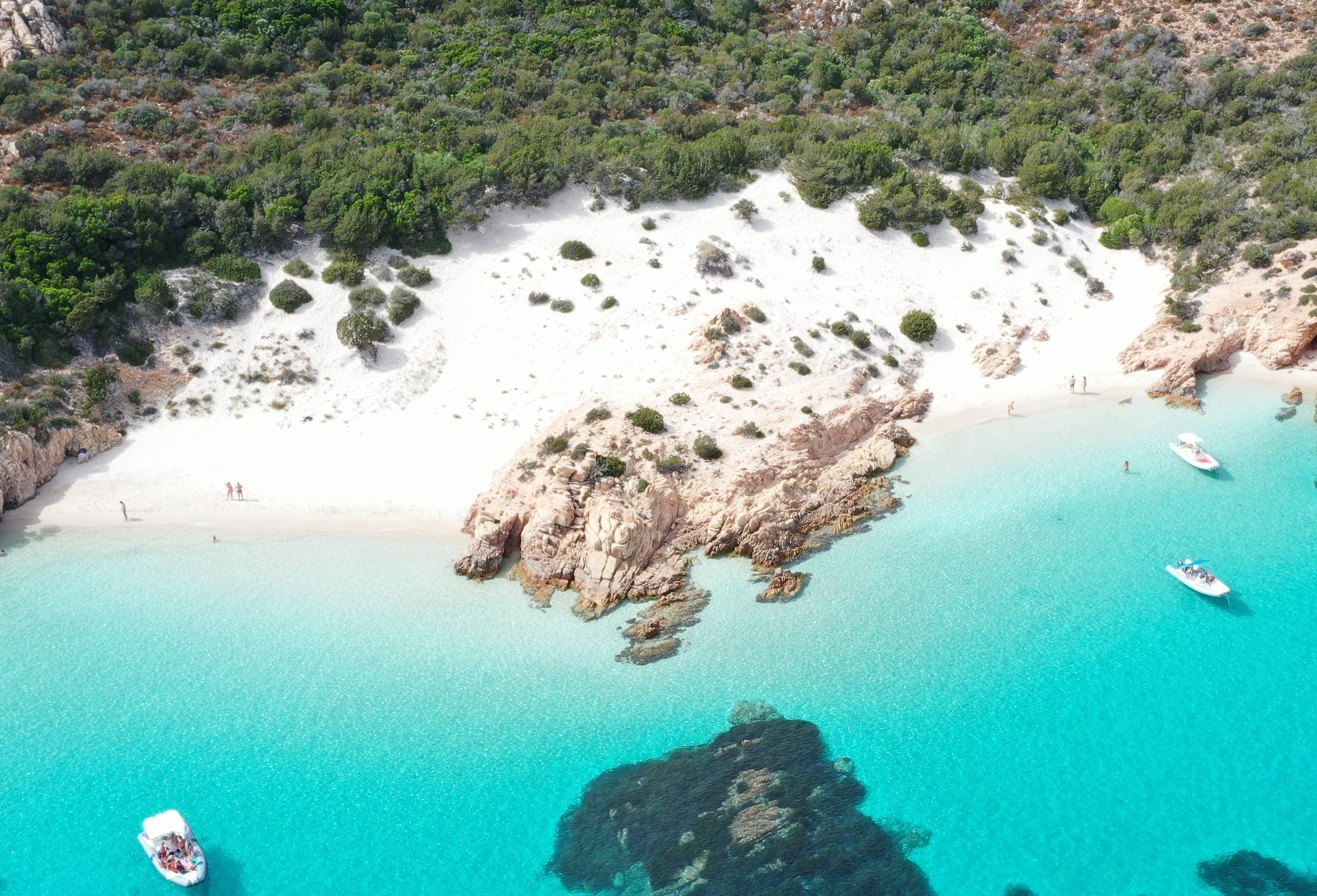 Immagine di una costa con spiaggia di sabbia bianca, scogliere e mare azzurro con barche a vela. Sullo sfondo una vegetazione verdeggiante.