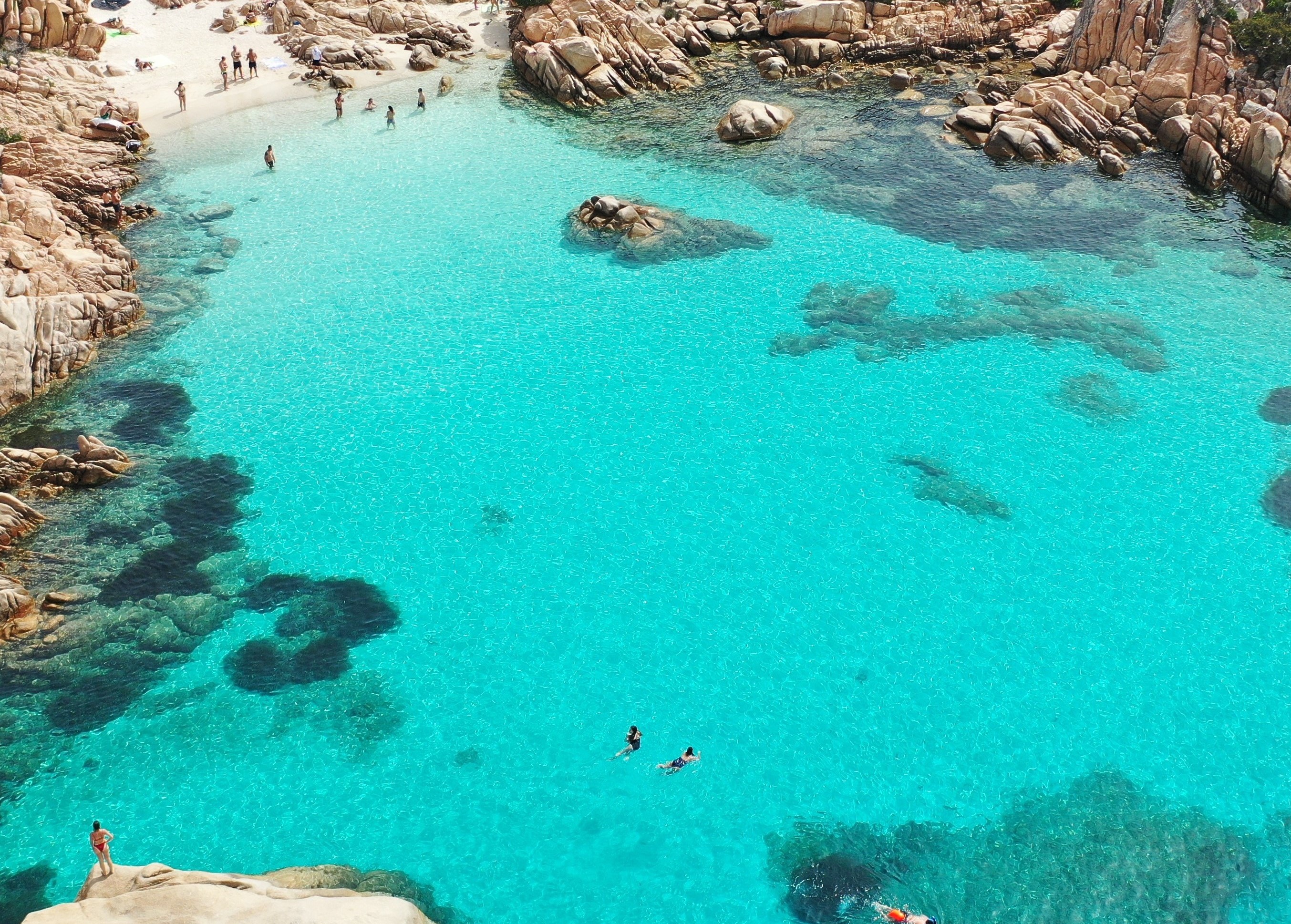 Immagine di un gruppo di persone che nuota in acque cristalline di un mare blu, con rocce e scogliere sullo sfondo, durante un tramonto.