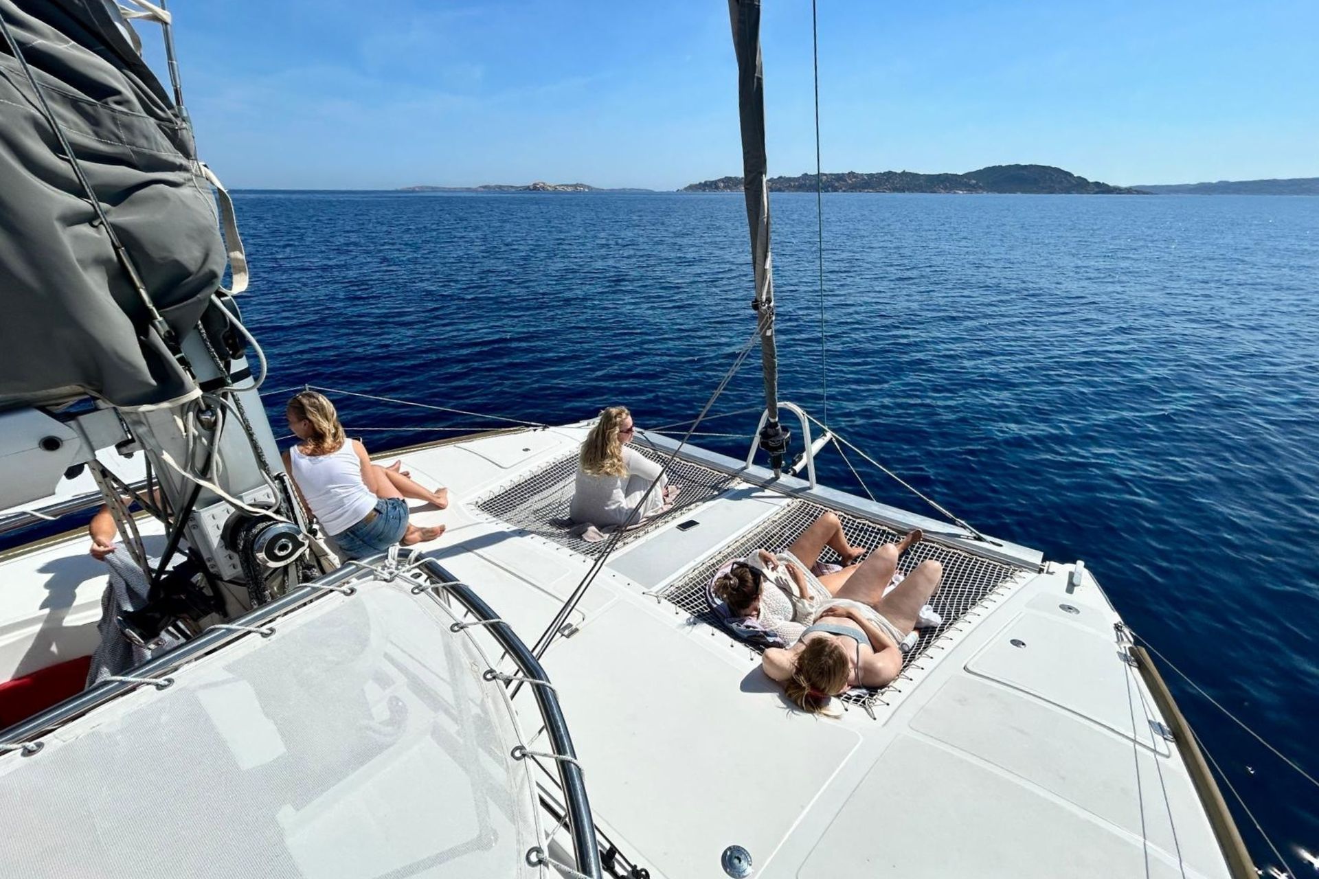 Drei Frauen genießen die Sonne an Bord eines Katamarans im Archipel von La Maddalena, mit kristallklarem Meer und Inseln im Hintergrund.