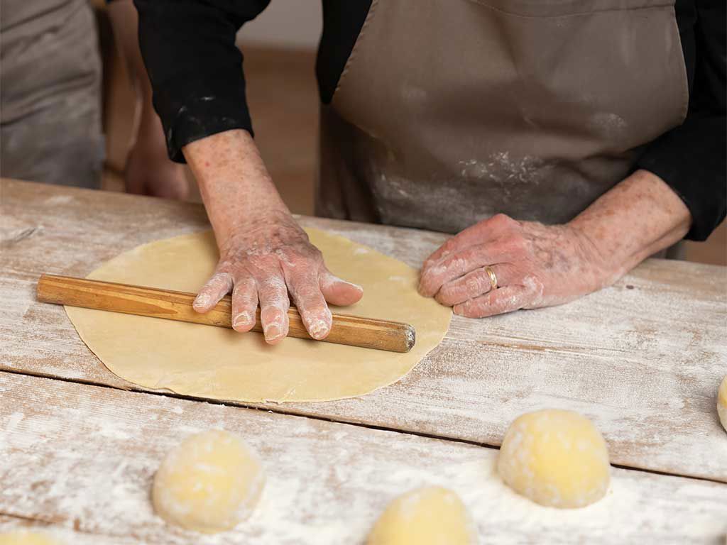 Una persona mayor prepara pasta fresca con un rodillo sobre una mesa de madera