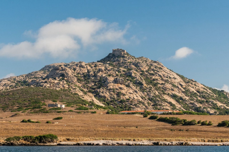 A rocky mountain with a fortress on the top and a desert landscape under a blue sky with some clouds.