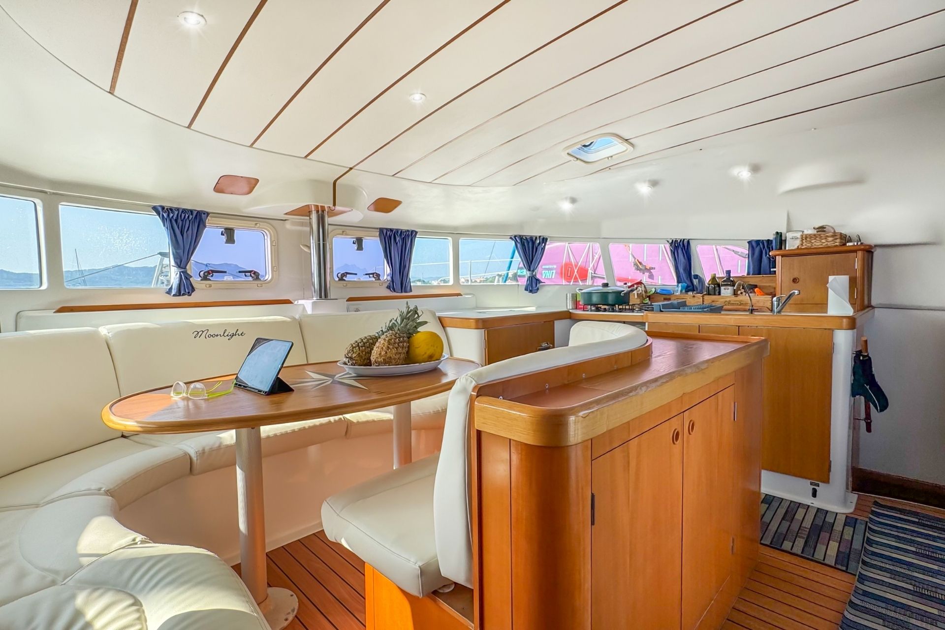 Interior de un catamarán con mesa, fruta, portátil y vista al mar. Se pueden ver ventanas con cortinas azules y una cocina con electrodomésticos y una cesta. El ambiente parece luminoso y acogedor.
