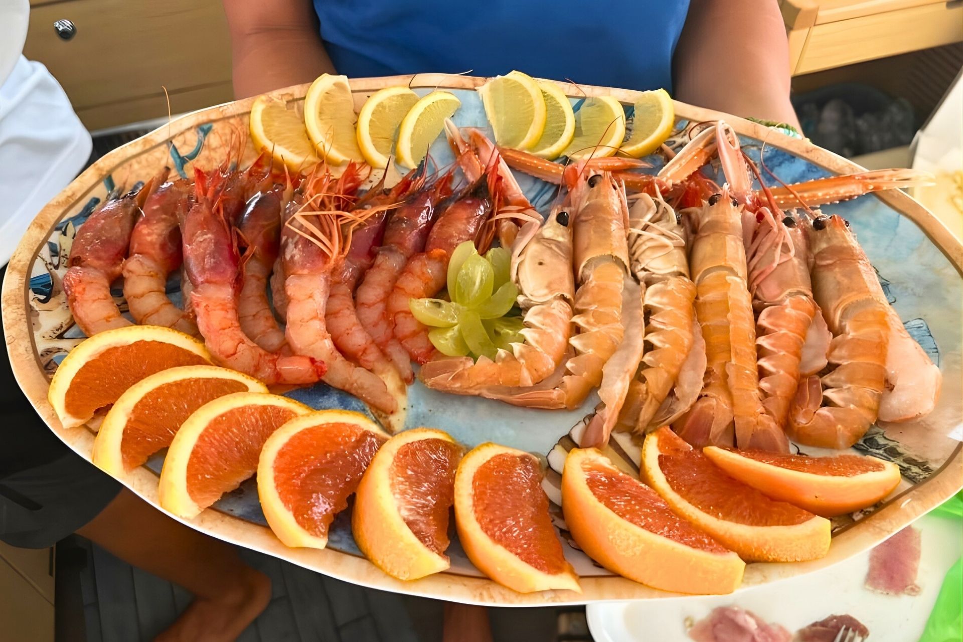Persona que sostiene un plato con camarones y naranjas en rodajas, probablemente a bordo de un catamarán.