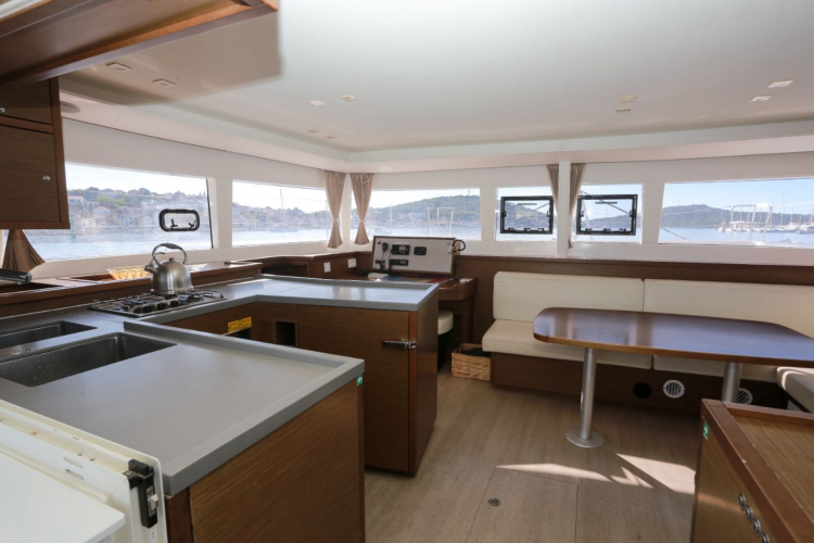 Interior of a catamaran with kitchen, table and sofa, windows with sea view and surrounding landscape visible from the outside.