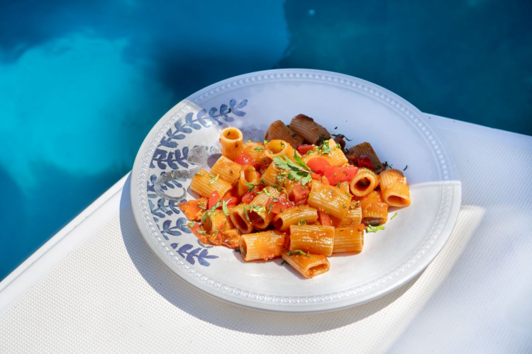 Pasta with tomato and basil served on a decorated plate, with the background of a swimming pool and blue water.