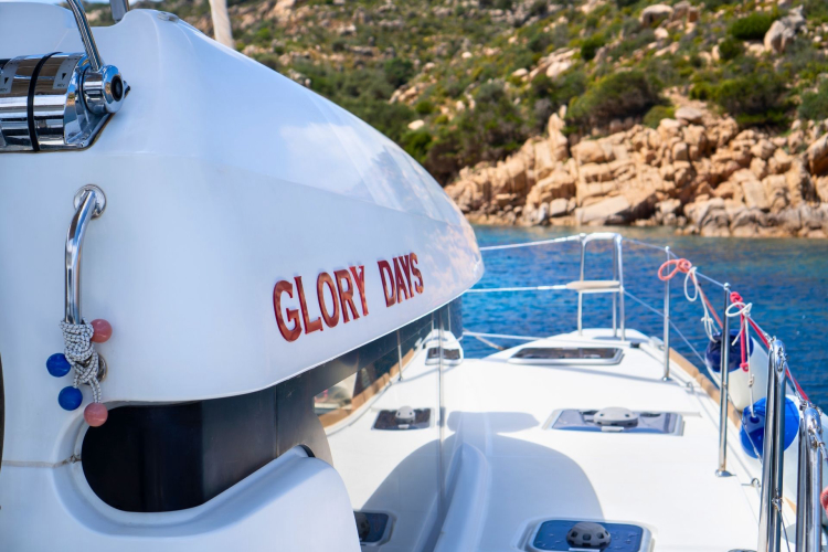 Luxury catamaran sailing with Glory Days written on the hull, a rocky coast with vegetation and calm sea in the background