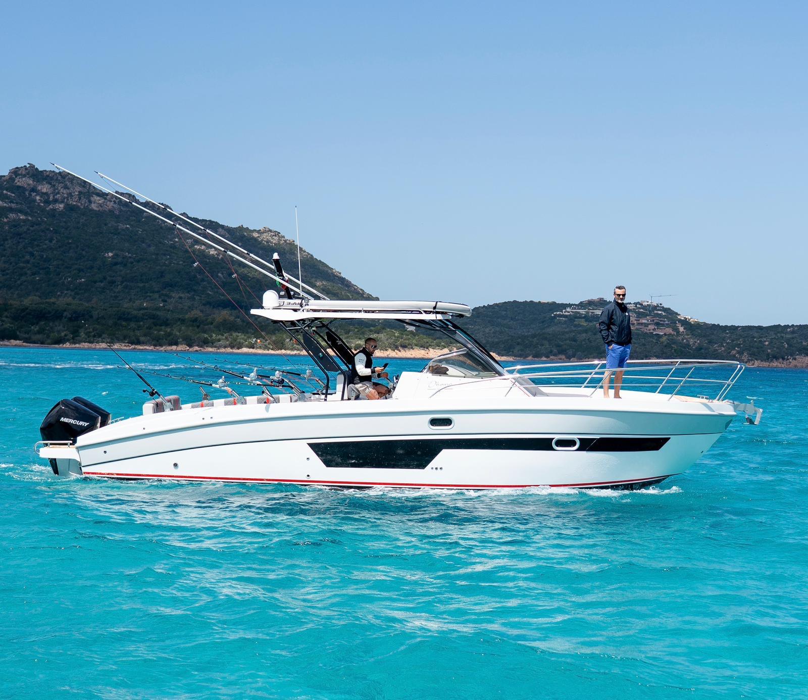 Dos personas en un barco pesquero, con montañas de fondo y mar azul turquesa.