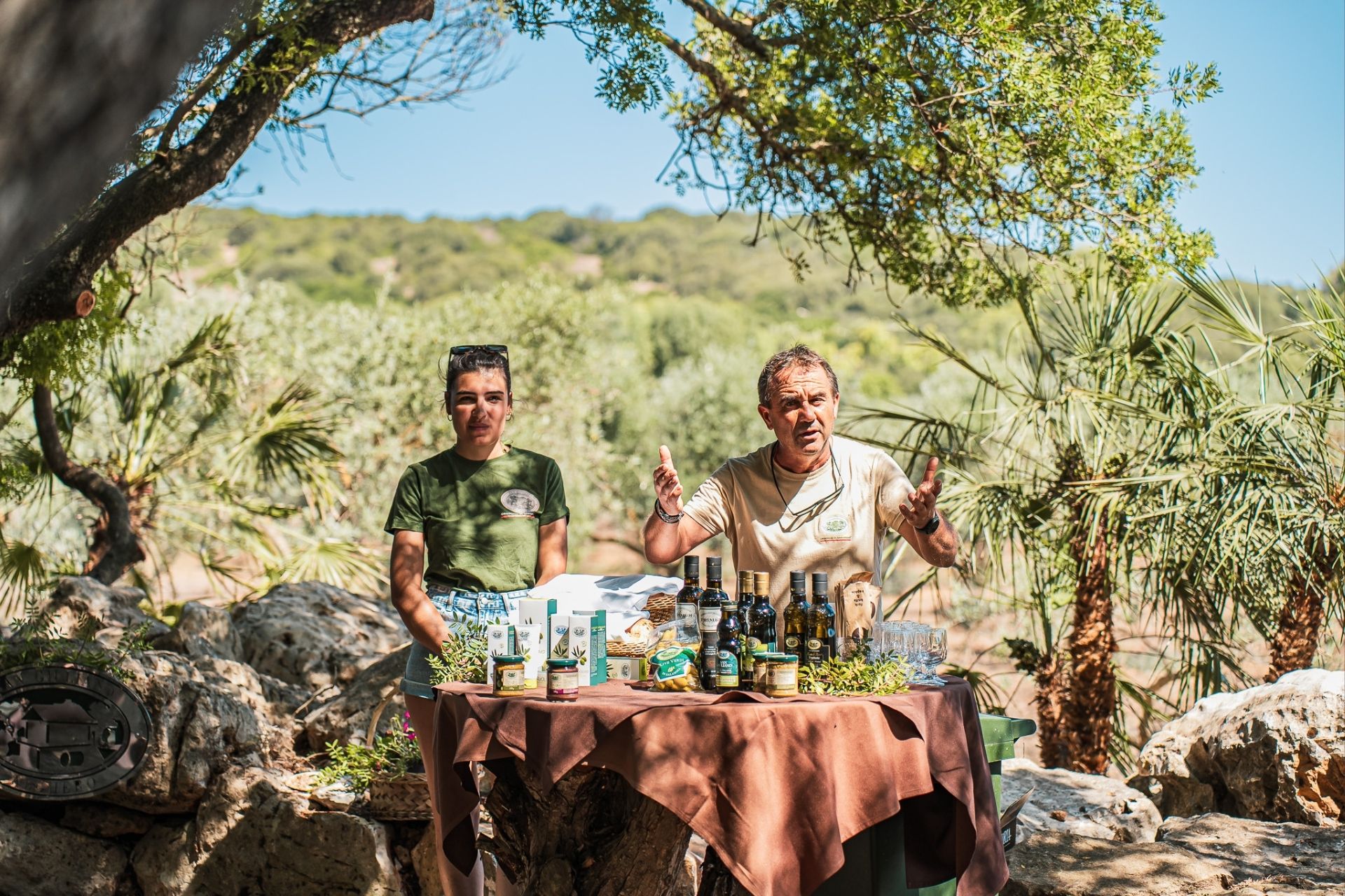 Dos personas en un olivar con una mesa llena de botellas de aceite y otros productos alimenticios