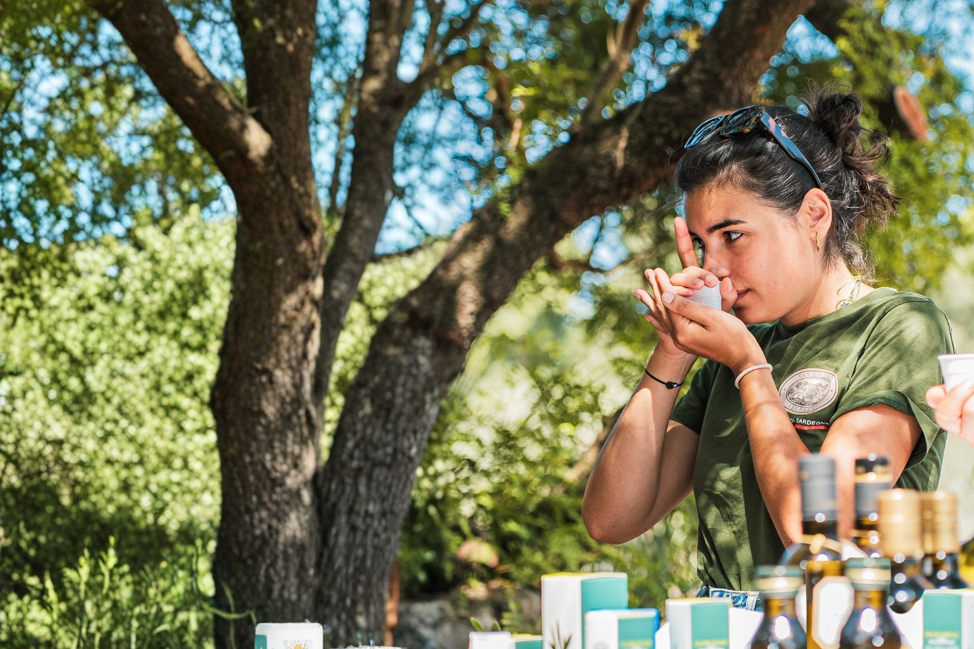 Una joven prueba aceite fresco en un olivar centenario de Alghero, Cerdeña, rodeada de botellas de aceite y otros productos locales.
