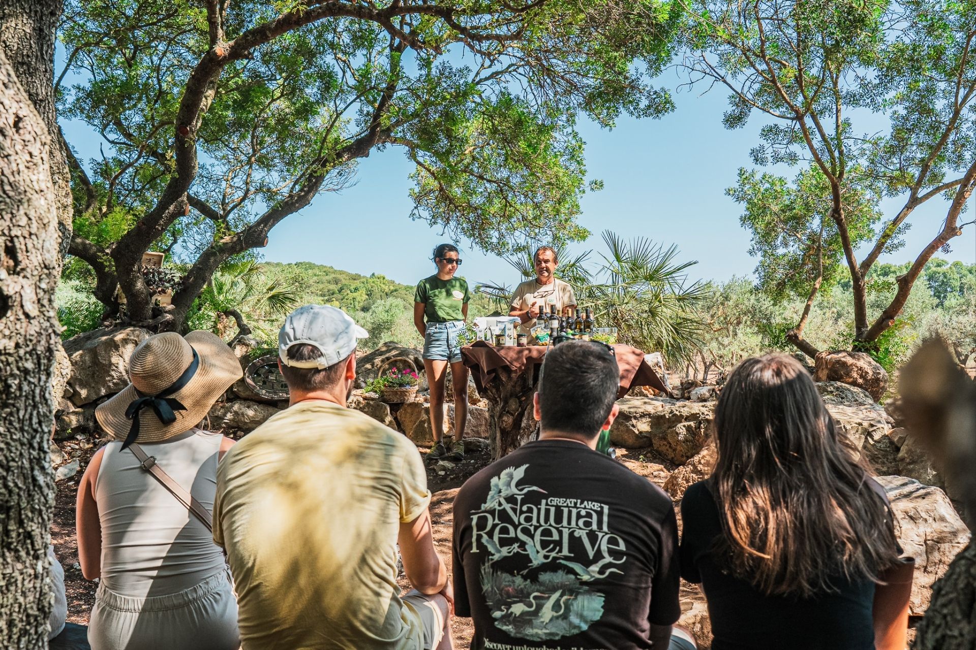 Grupo de personas sentadas al aire libre en un olivar con un guía hablando frente a una mesa con botellas de aceite