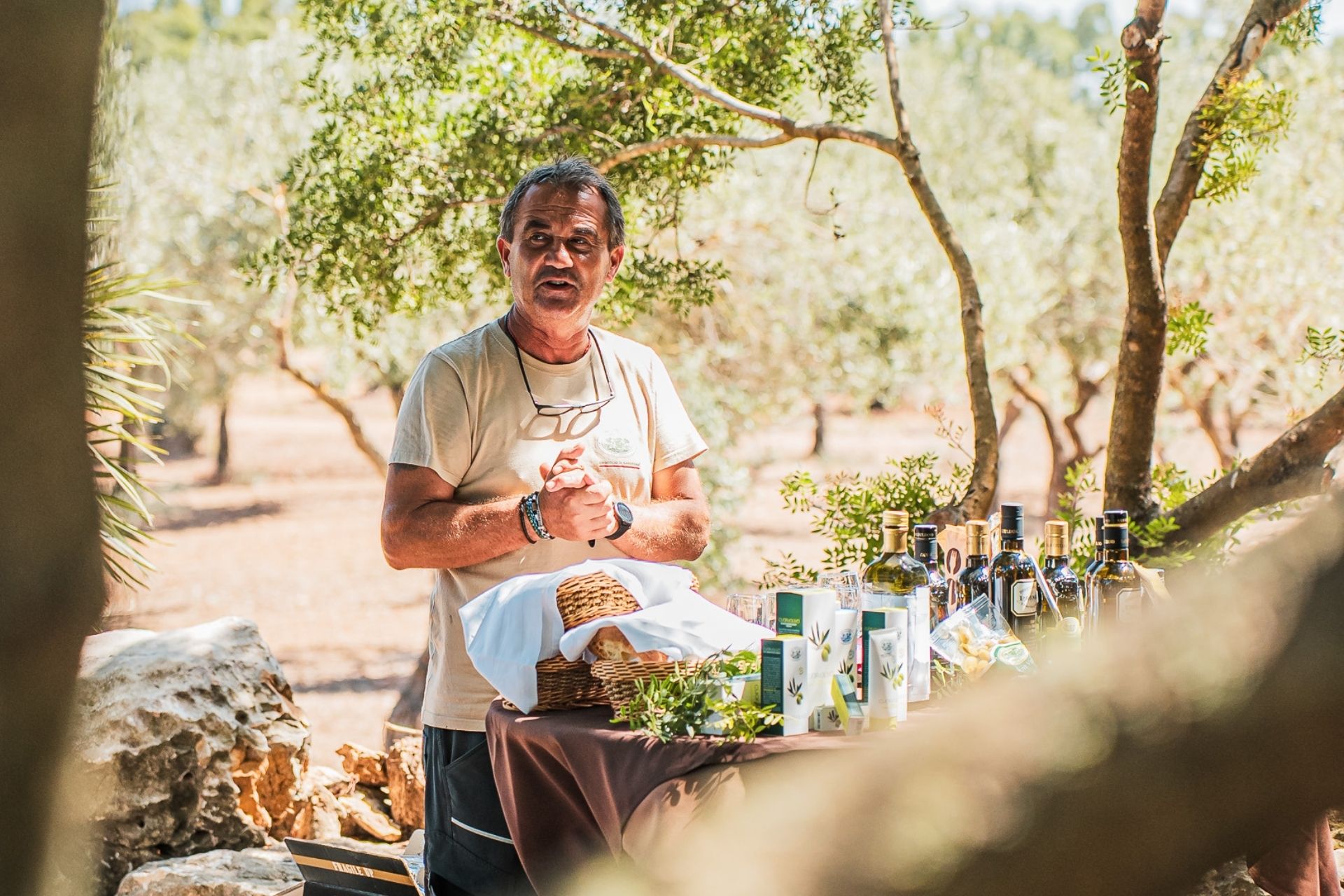 Un hombre de pie en un olivar con una mesa con botellas de aceite y otros productos locales.