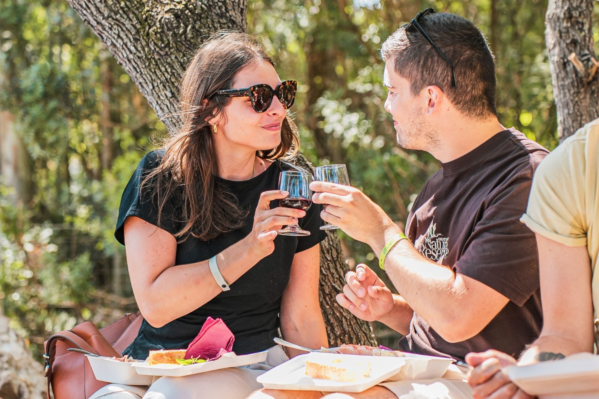 Dos personas sentadas bajo un árbol, una de las cuales sostiene una copa de vino y la otra un plato de comida.
