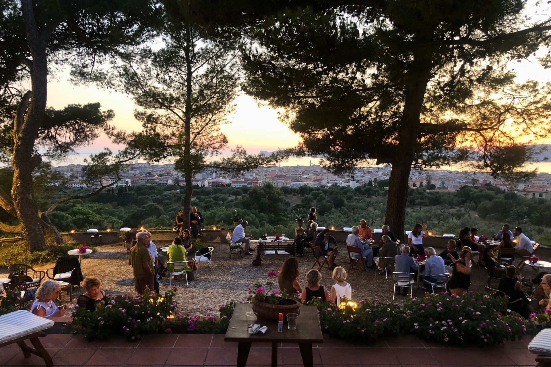 A group of people sitting around tables in a green area with a panoramic view of Alghero