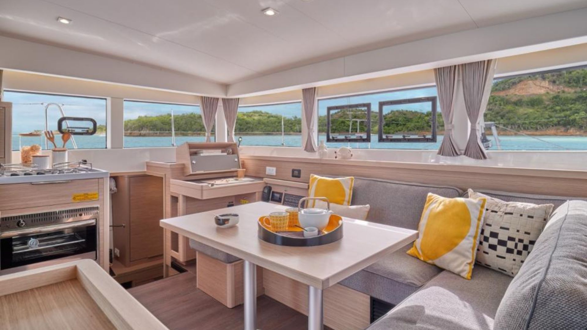 Intérieur d'un catamaran avec table, canapés et cuisine, fenêtres panoramiques donnant sur la mer et les îles