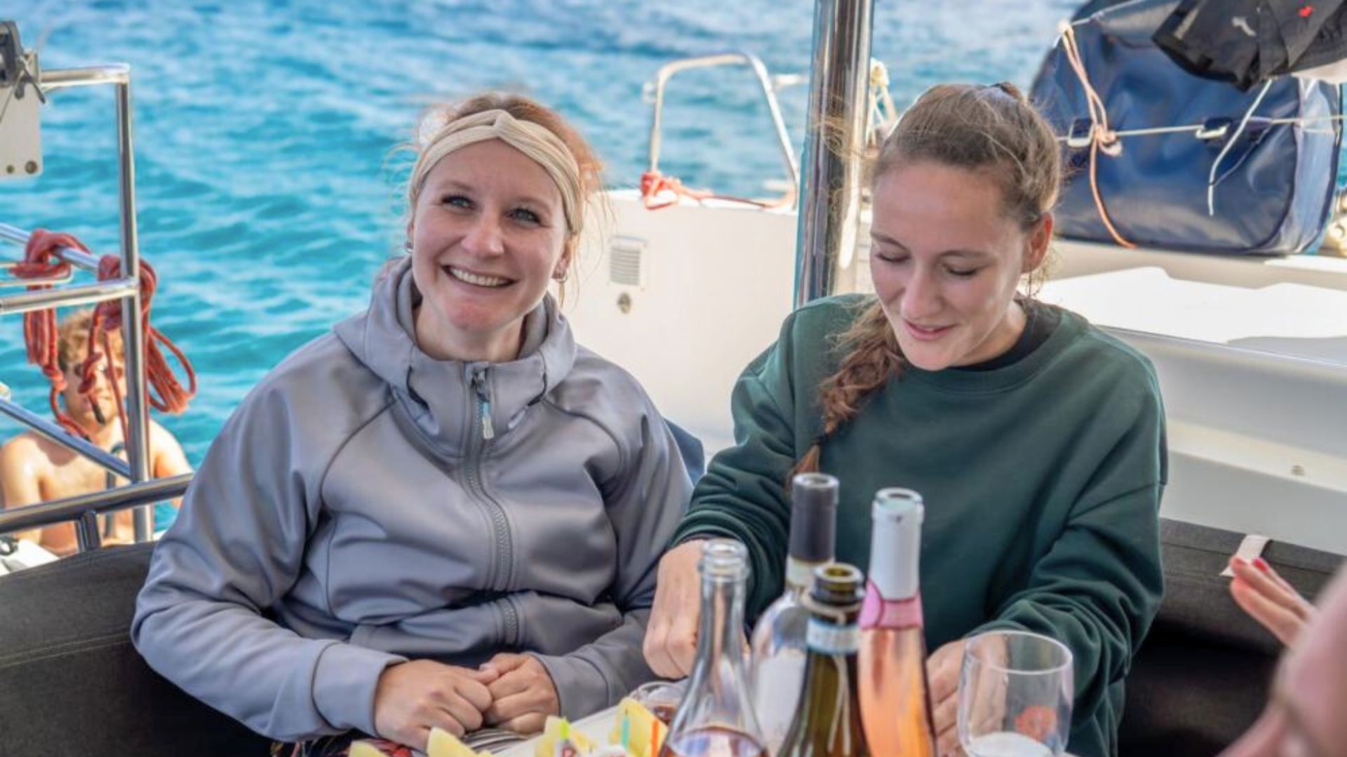Deux femmes souriantes assises sur un catamaran, avec une table pleine de bouteilles