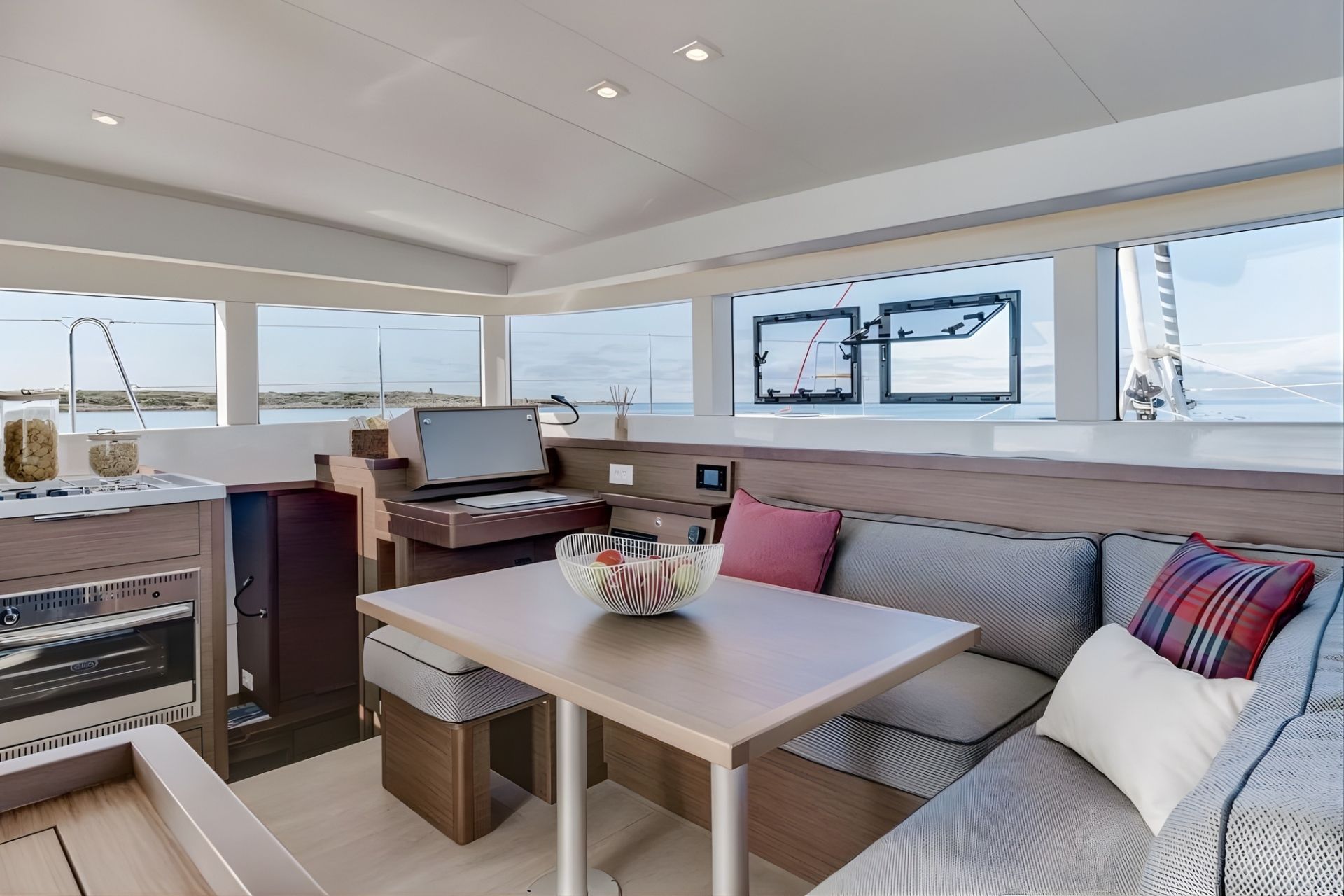 Interior de un catamarán con mesa, sofá y monitor, vista de un mar de aguas cristalinas e islas al fondo