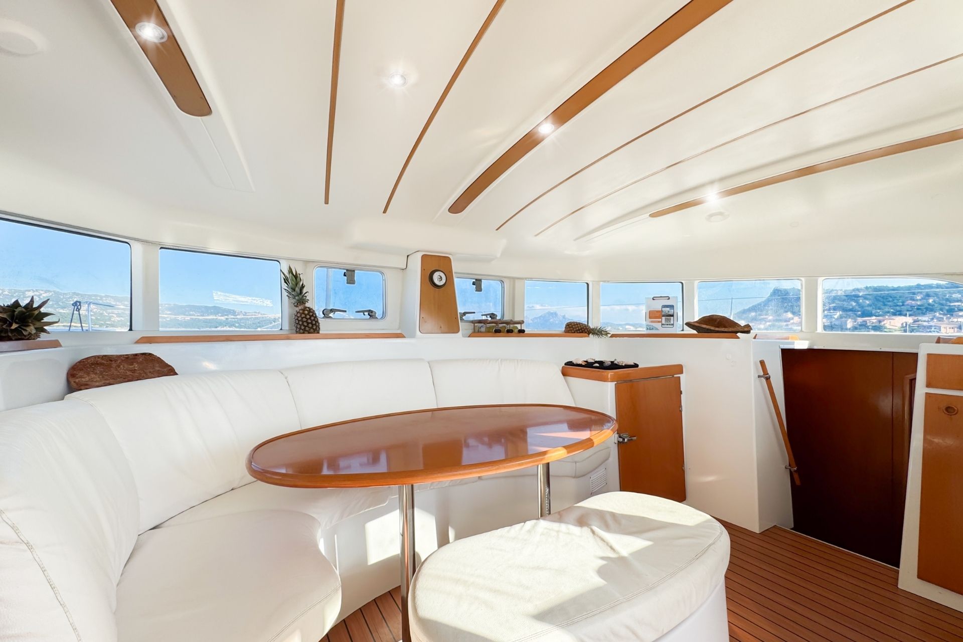 Interior of a catamaran with white sofa, round table, plants and panoramic view of islands and crystal clear sea.