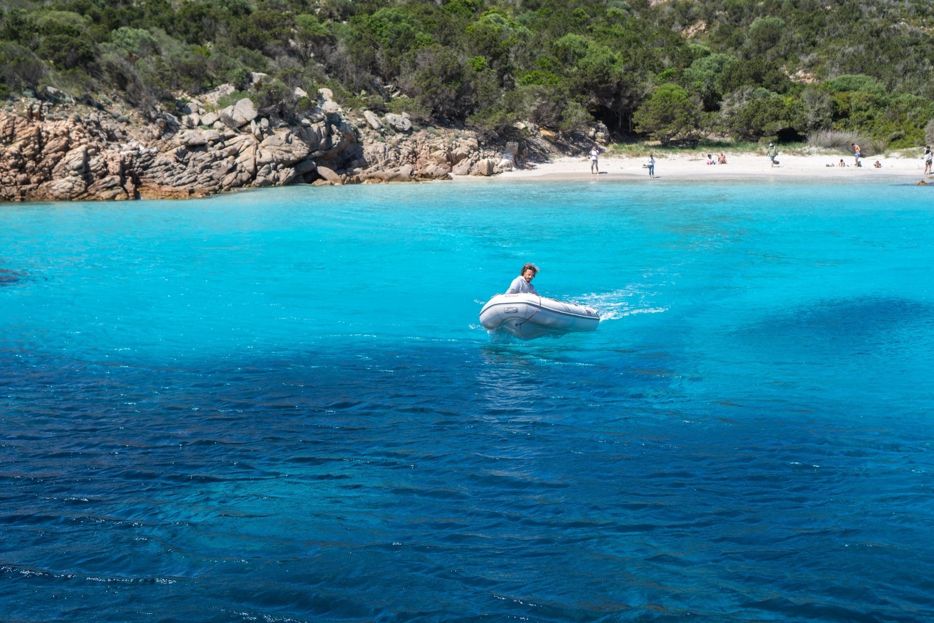 A man in a dinghy navigates crystal blue waters, with a rocky beach and people in the background.
