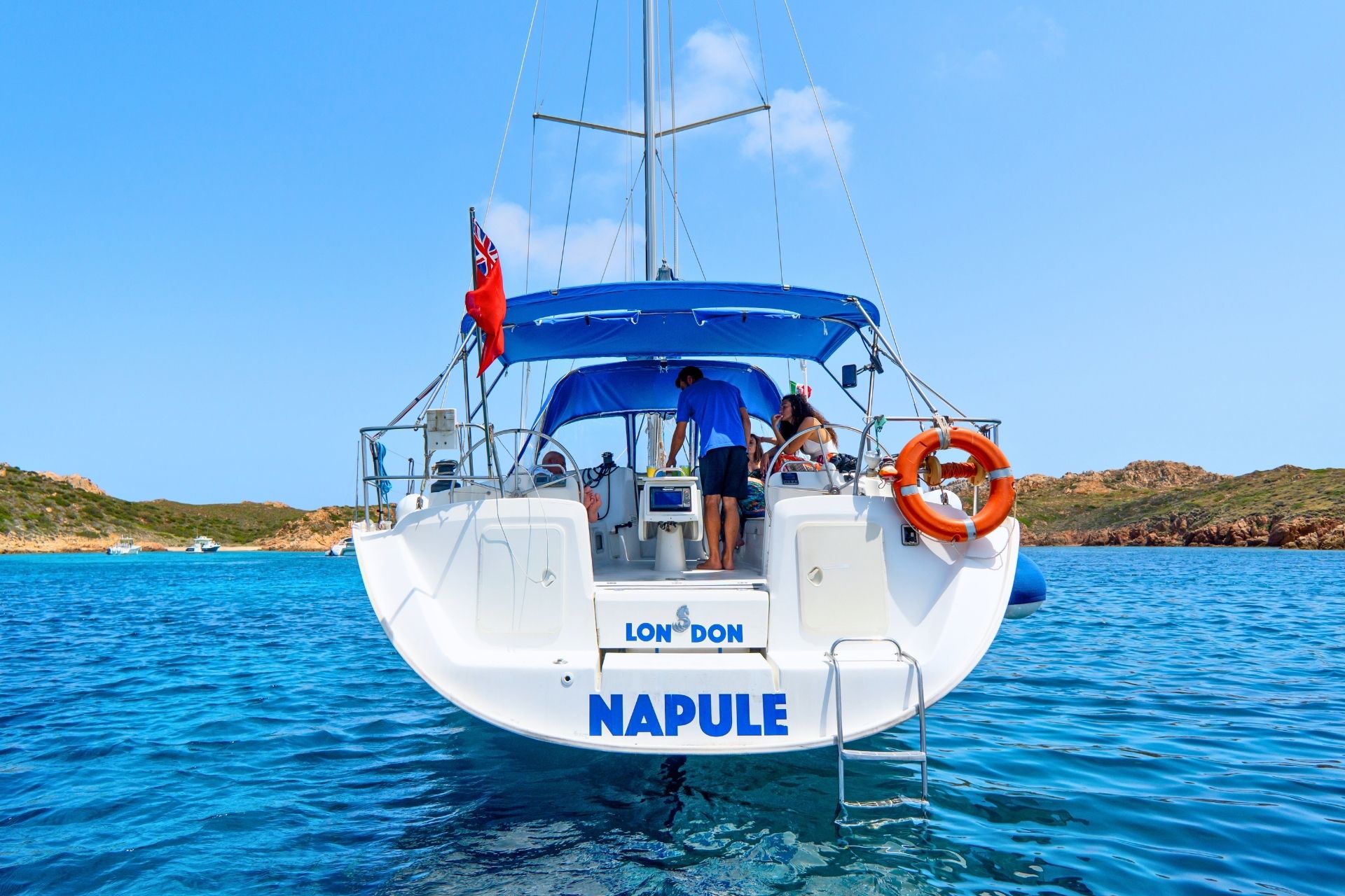 Un bateau avec des personnes à bord navigue dans des eaux cristallines, avec une île en arrière-plan. Le bateau s'appelle Napoule.