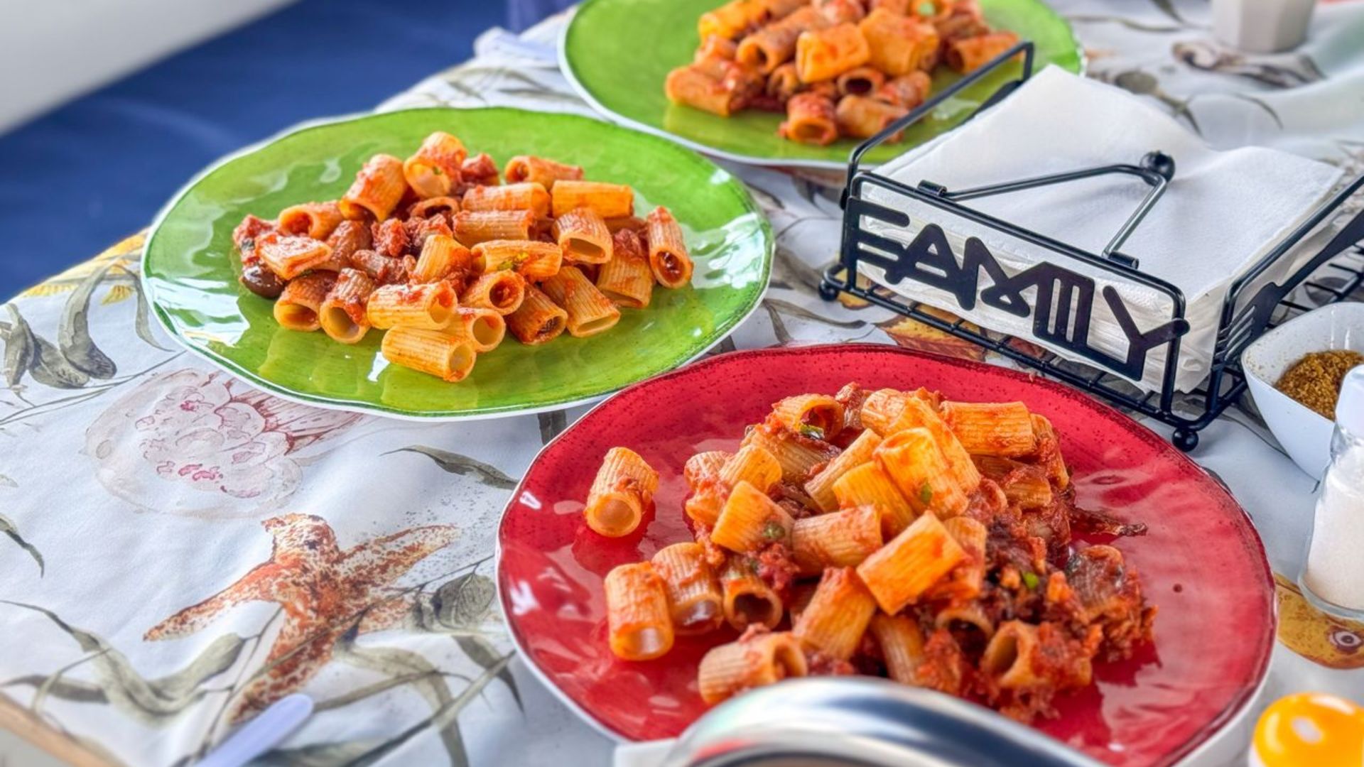 Tres platos de pasta servidos en platos coloridos sobre una mesa con un mantel floral