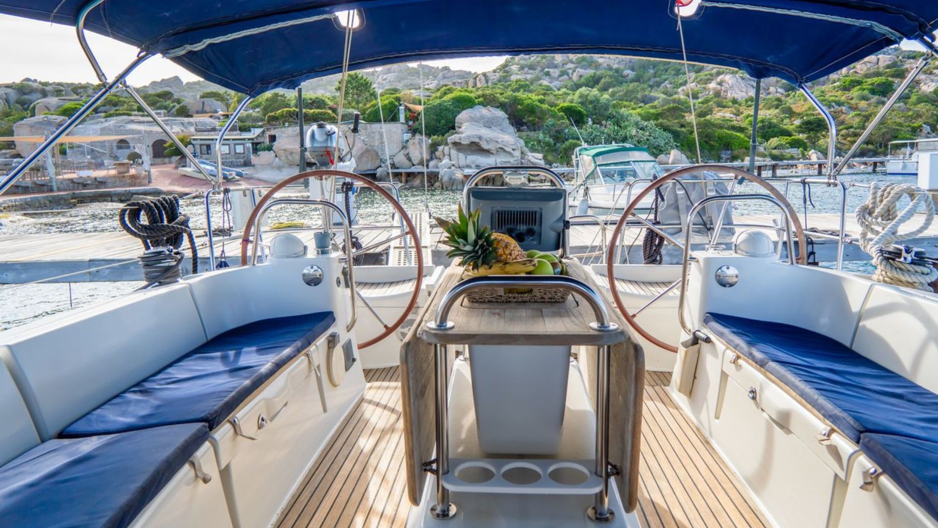 Interior de un barco con asientos azules, una mesa con fruta y un puerto al fondo