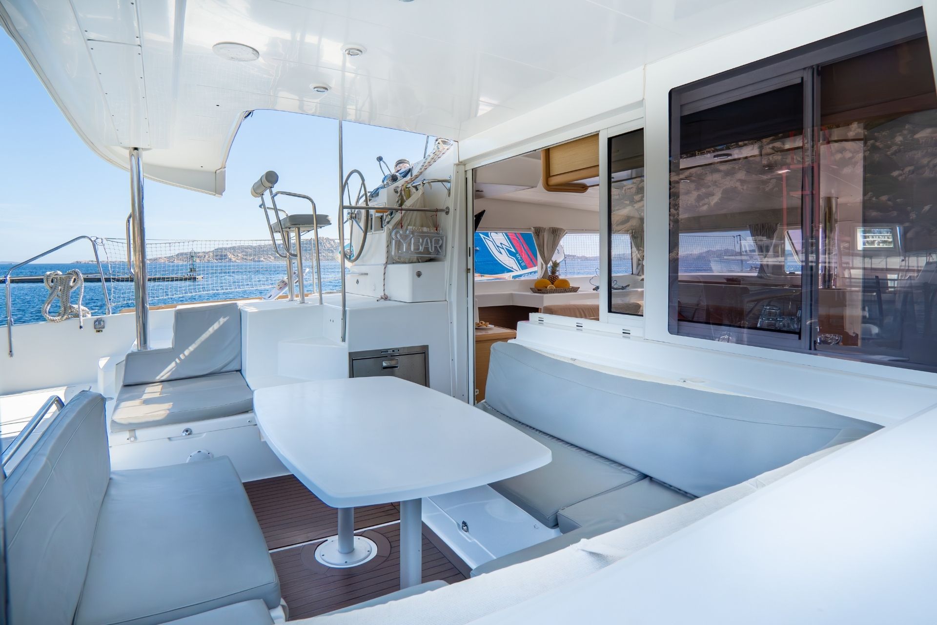 Interior of a catamaran with table and seats, view of the sea and islands in the background, with a sign that says 'BAR'.