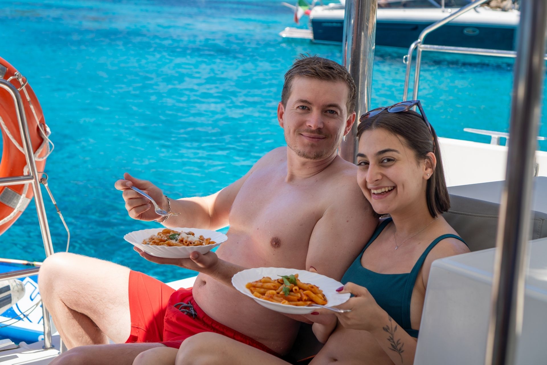Couple sitting on a catamaran with food in their hands, in the background an island with blue sea