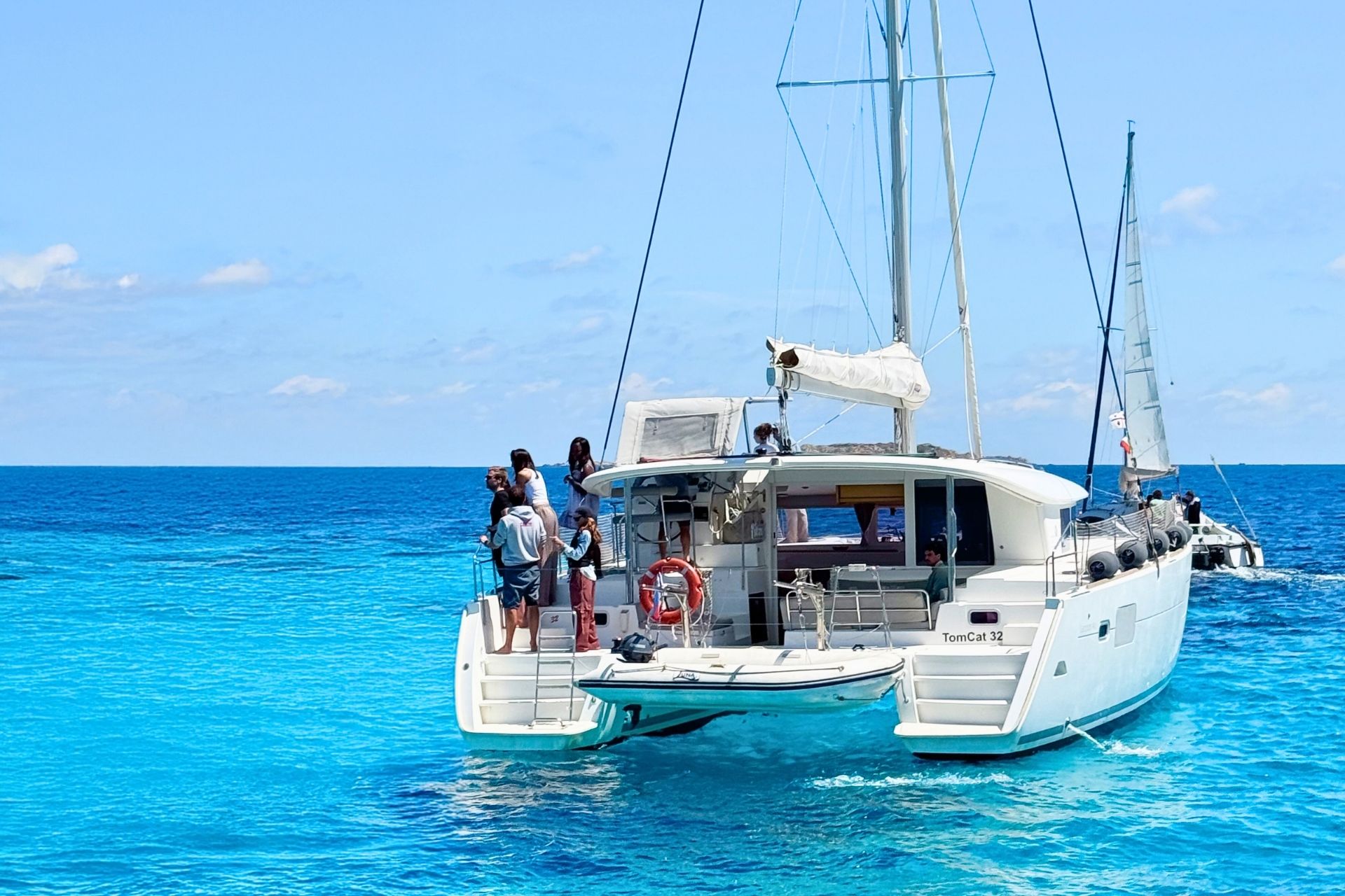 A group of people enjoy a sunny day aboard a catamaran in the archipelago of La Maddalena, with crystal clear waters and paradisiacal islands in the background.