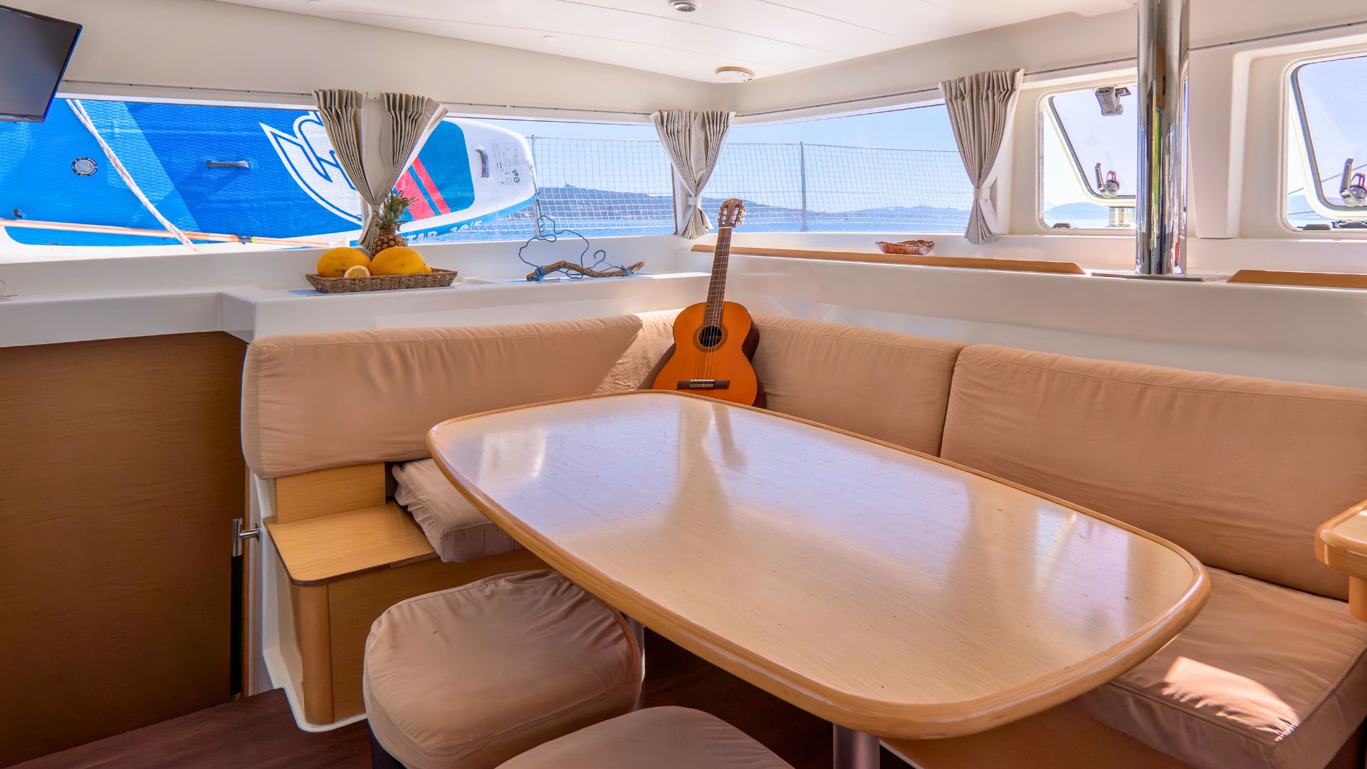 Interior de un catamarán con mesa, sofá y ventanas panorámicas, con guitarra y fruta a bordo. Fuera de la ventana se puede ver una isla y otro barco