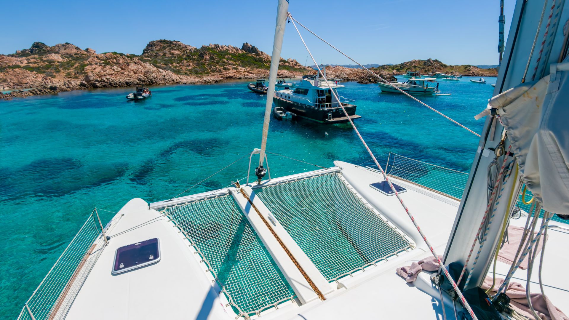 Imagen de un catamarán y otros barcos en una bahía con aguas cristalinas y rocas