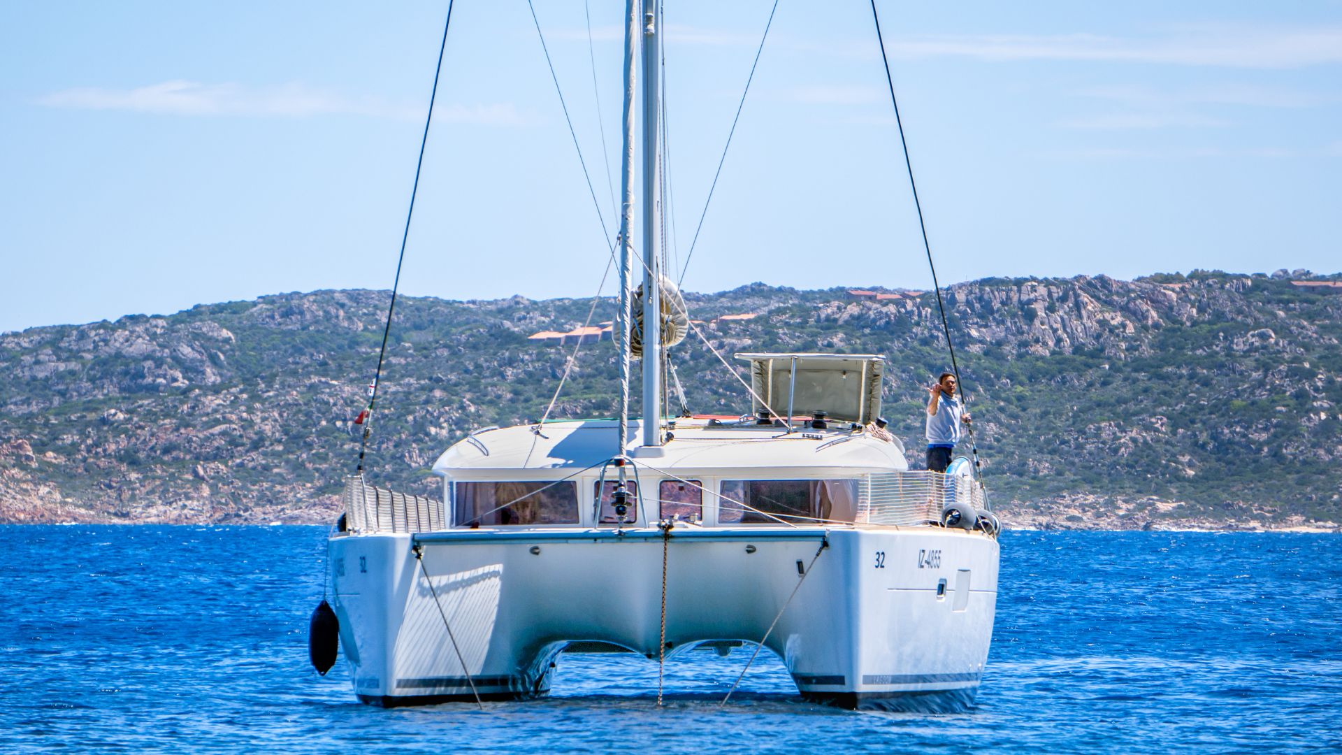 Un catamarán blanco navega hacia el mar con un hombre de pie a bordo que saluda