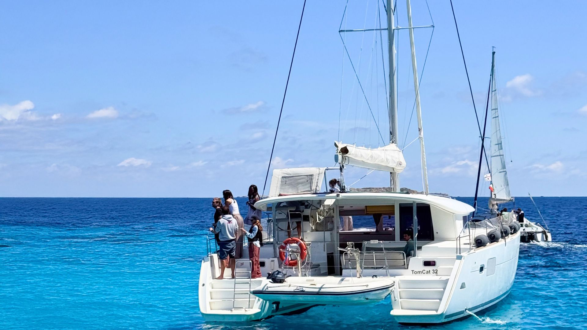 Un grupo de personas de pie en un catamarán de vela con mar en calma y cielo azul