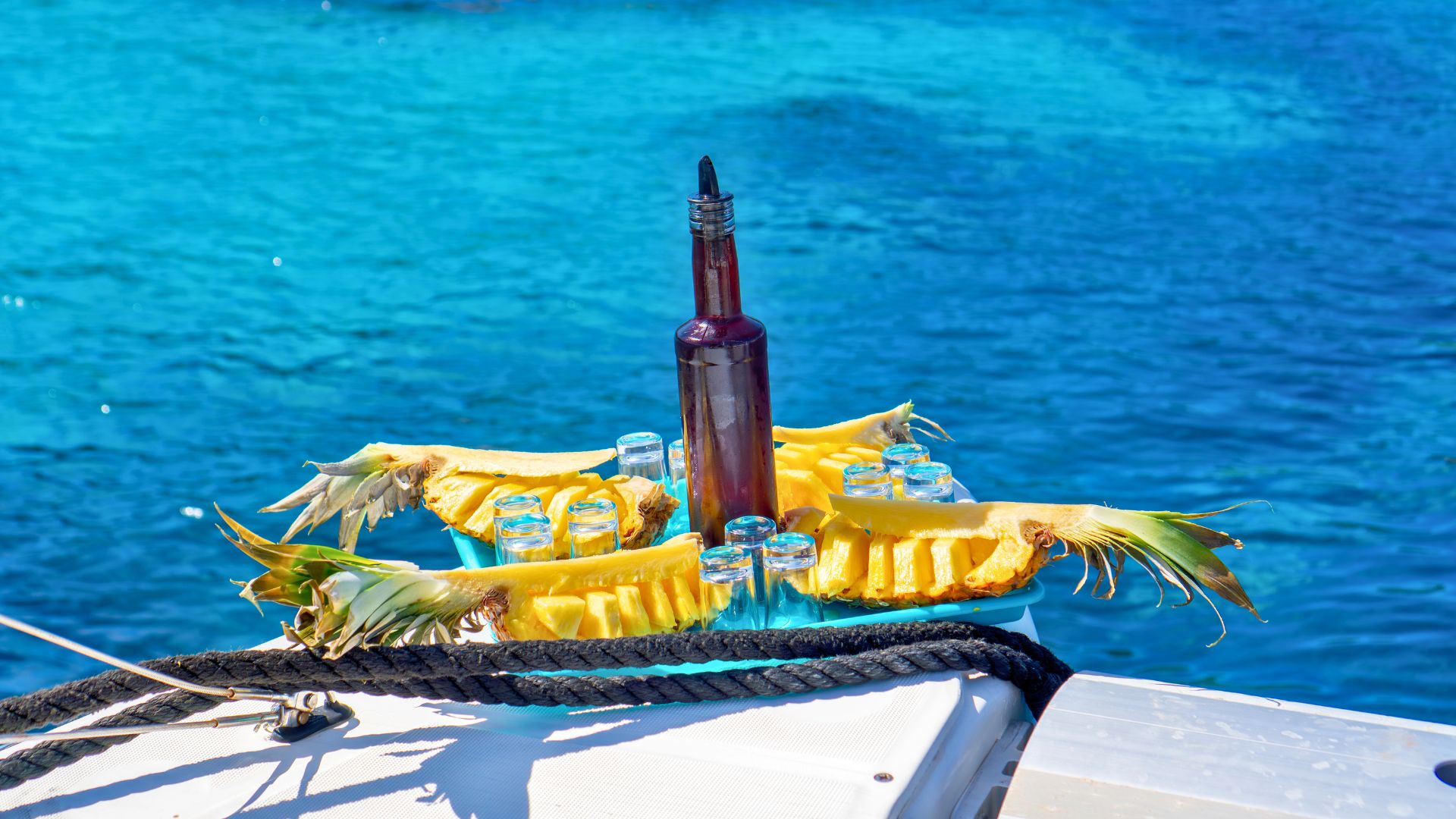 Catamarán con botella y vasos en una bandeja, rodeado de piña, con el fondo del mar azul