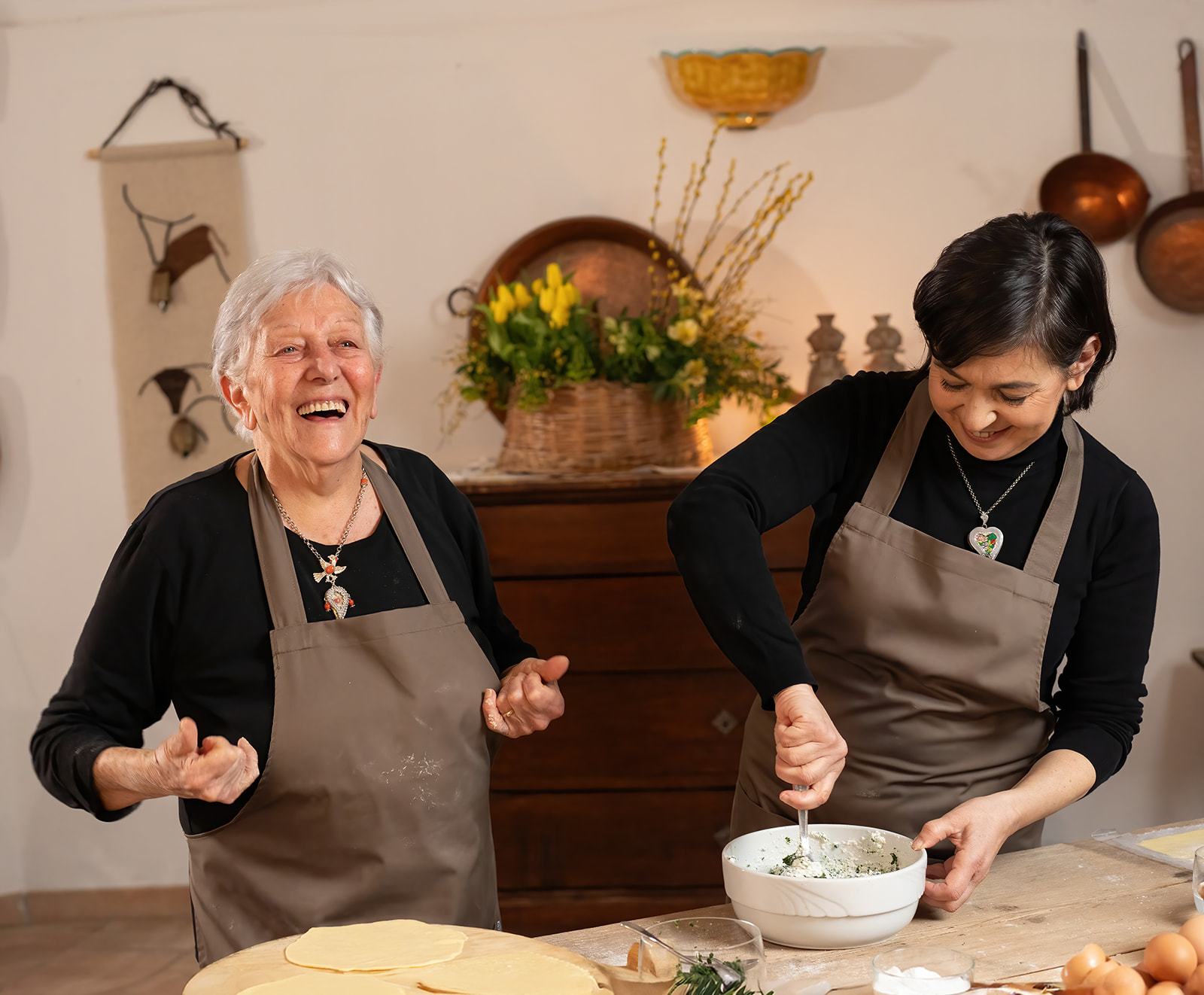 Dos mujeres en un entorno rústico, probablemente un taller de cocina, se preparan para cocinar. Una de ellas lleva un delantal y sonríe, mientras que la otra mezcla un ingrediente en un bol