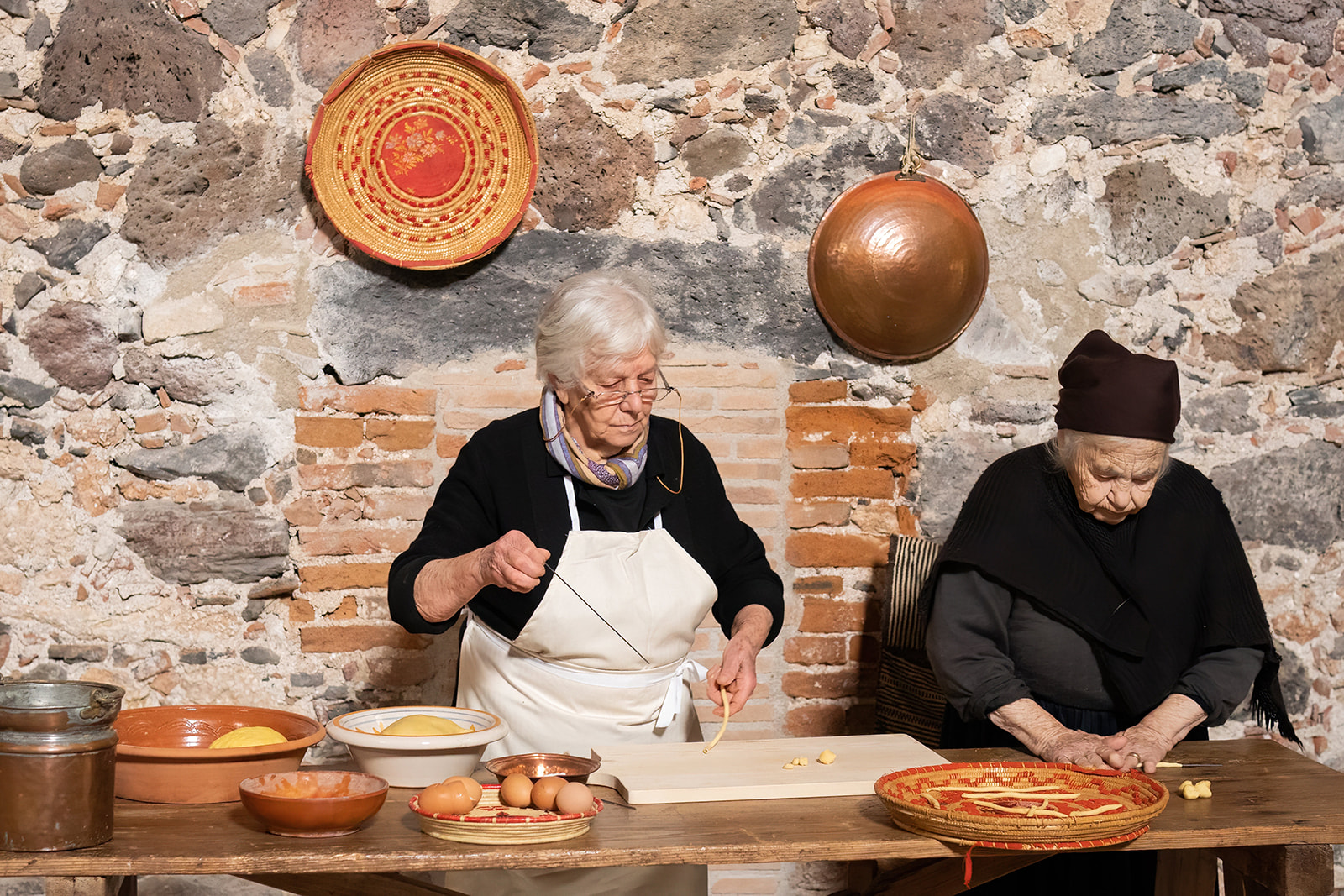 Dos ancianas preparan postres típicos de Cerdeña en una cocina tradicional, rodeadas de ingredientes frescos y utensilios de cocina