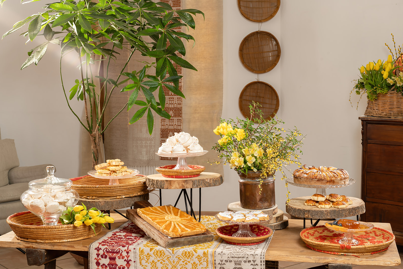 Una mesa con dulces típicos de Cerdeña en platos de madera y dulces en bandejas de vidrio, con flores amarillas en macetas