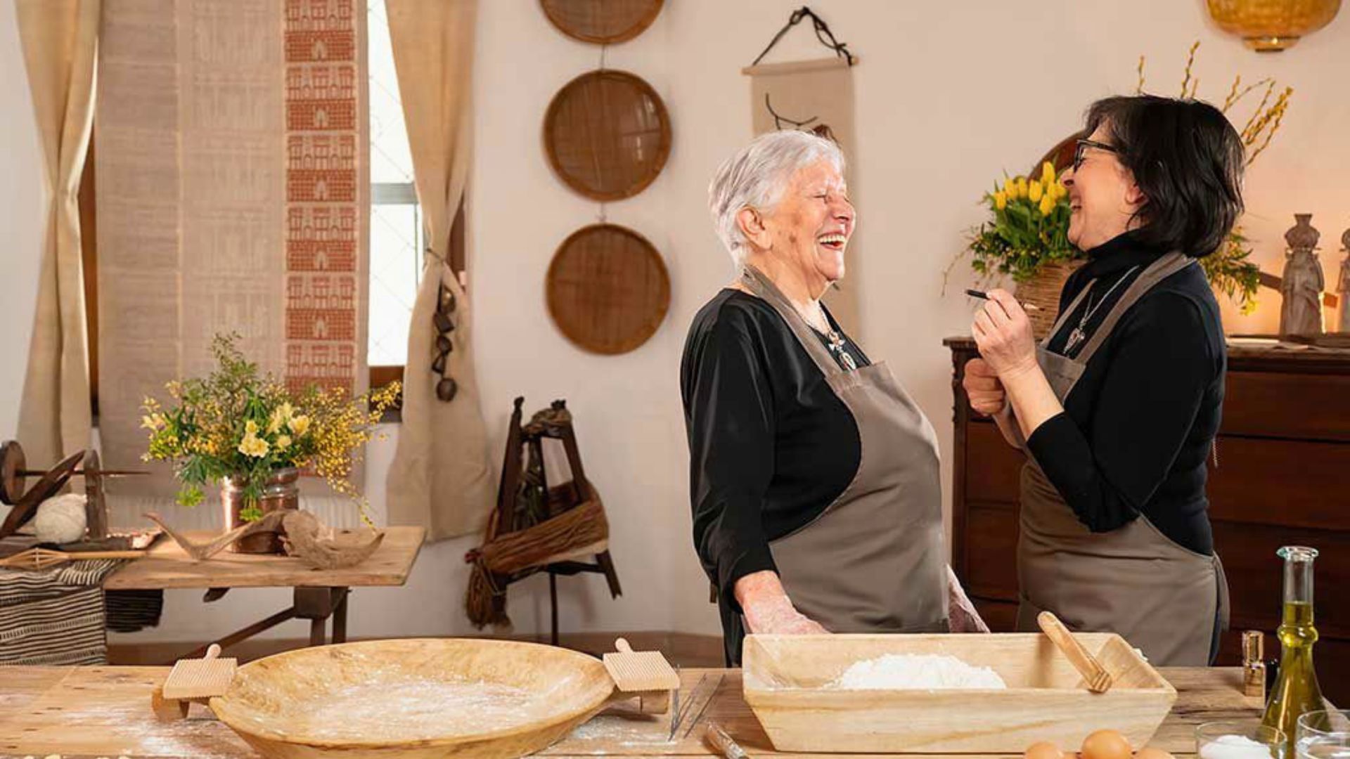 Due donne con grembiuli in una cucina, una che sorride e l'altra che scrive, con ingredienti per la pizza e un ambiente accogliente
