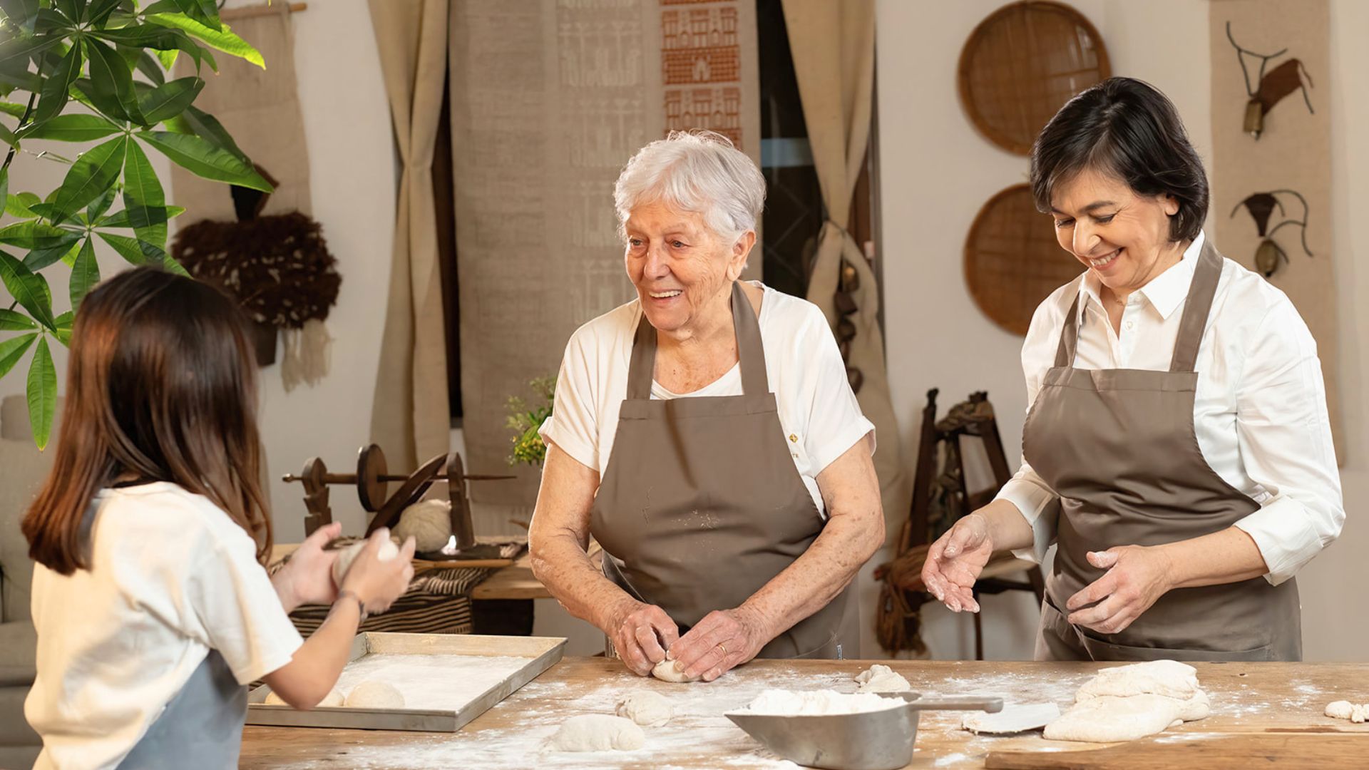 Tre persone, due donne e una bambina, lavorano insieme a un tavolo di legno, creando e preparando la pizza. Sullo sfondo sono presenti piante e altri oggetti decorativi