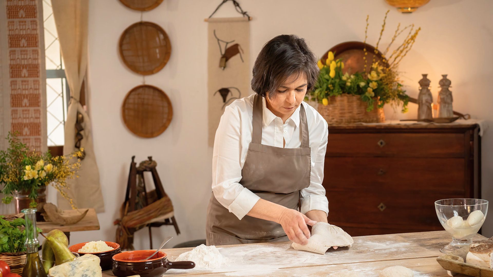 Una donna prepara impasti di pizza in una cucina tradizionale, circondata da ingredienti e utensili da cucina. Sullo sfondo, ci sono fiori e altri oggetti decorativi