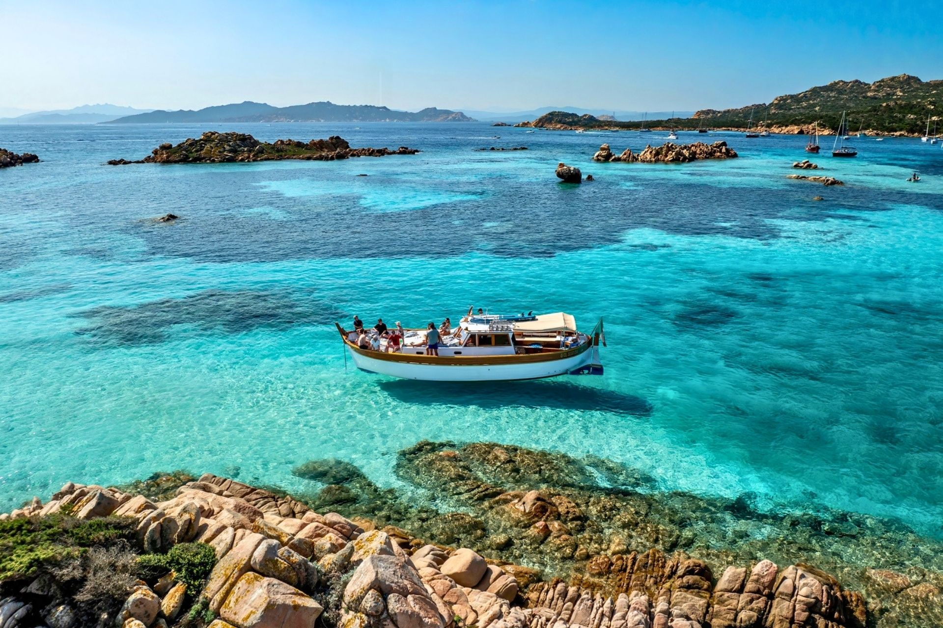 A vintage boat sails in turquoise waters with rocky islands in the background and a group of people on board