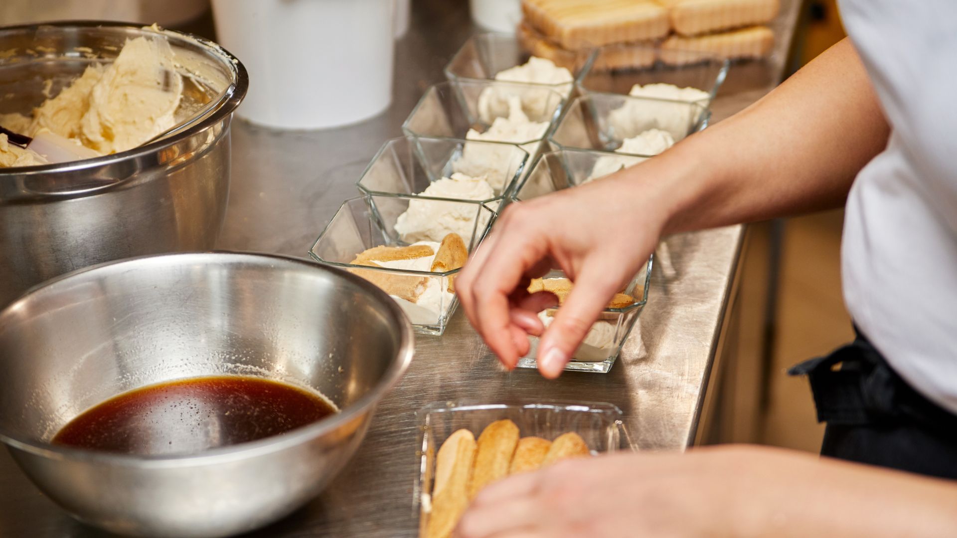 A person in the kitchen is preparing a tiramisu with fresh ingredients in a glass container