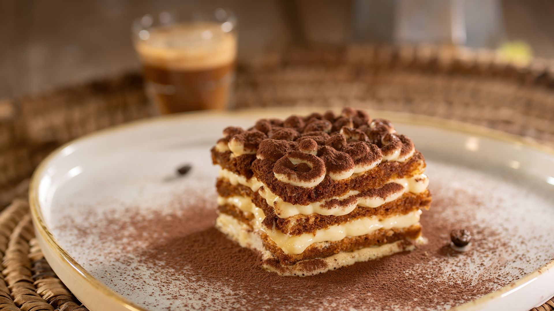 A sweet tiramisu on a plate with cocoa powder and a glass of chocolate drink in the background
